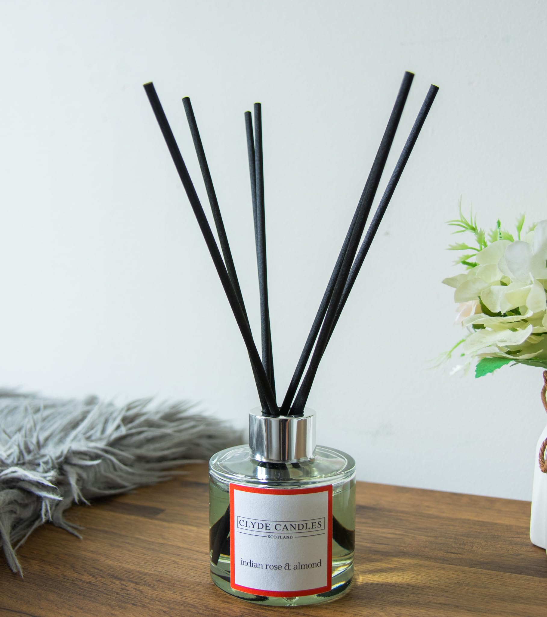 A Clyde Candles reed diffuser featuring Indian rose and almond scent, placed on a wooden surface, accompanied by a decorative plant and cozy rug.